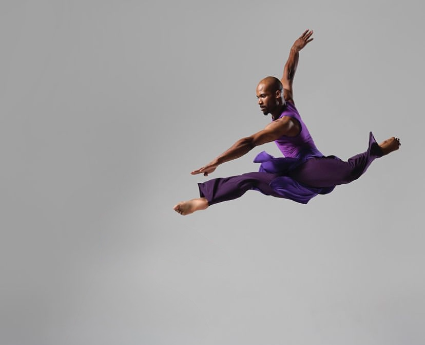A person in a purple outfit is mid-air performing a leap against a plain gray background.