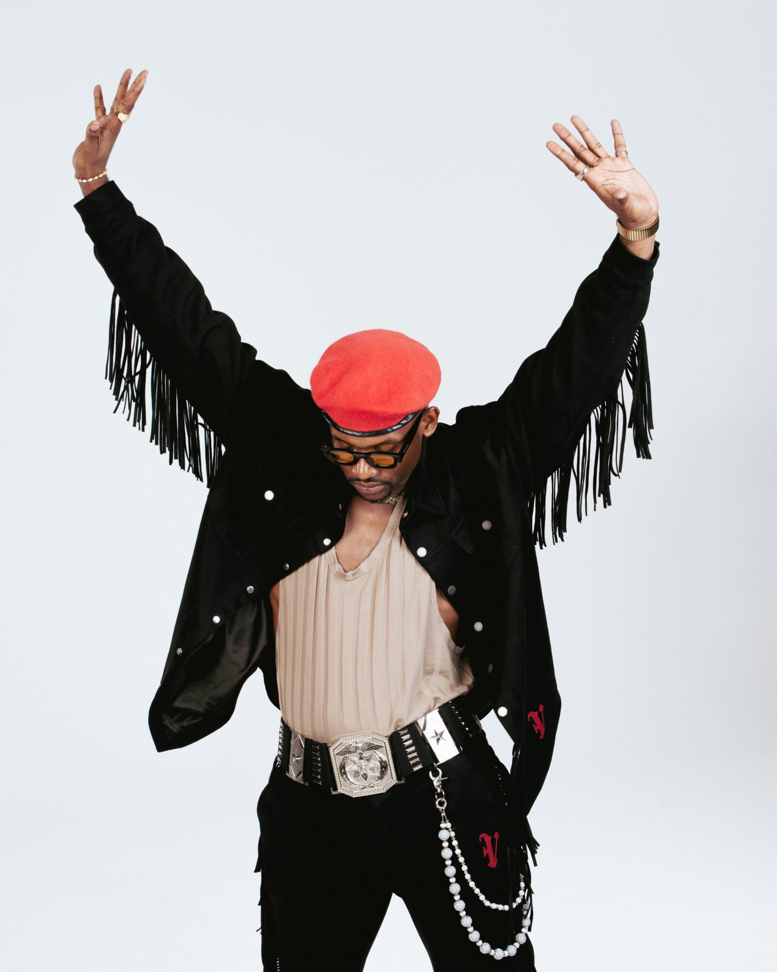 Person with arms raised, wearing a red hat, black fringed jacket, beige top, and accessories, against a light background.