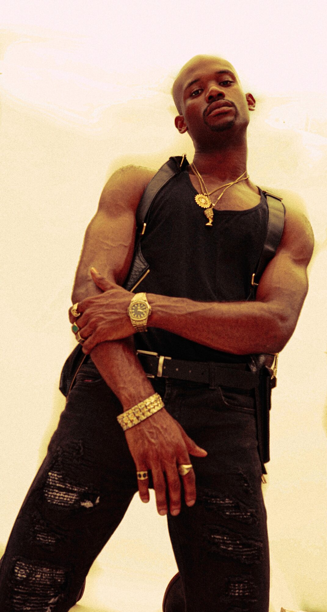 Man with muscular arms wearing black sleeveless shirt, gold jewelry, and distressed black jeans, standing against a plain background.