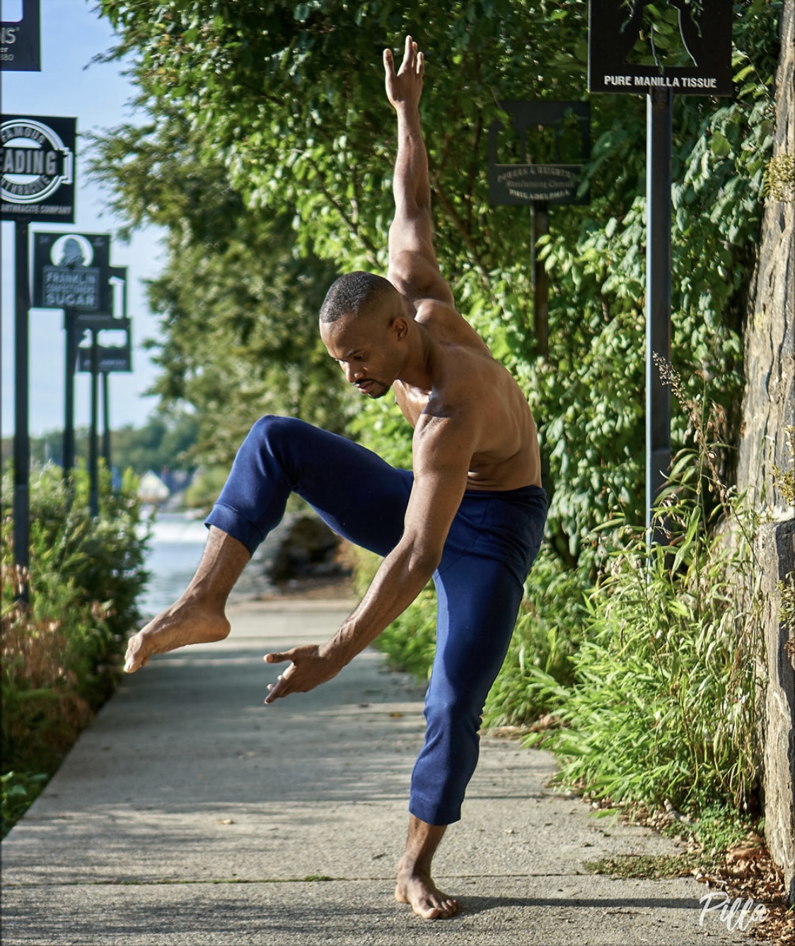 A shirtless man with a shaved head balances on one foot on a sidewalk, with one arm raised and the other extended downward.
