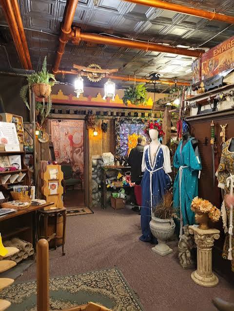 Interior of a vintage shop with mannequins dressed in historical costumes, shelves with books and decorative items, and plants.
