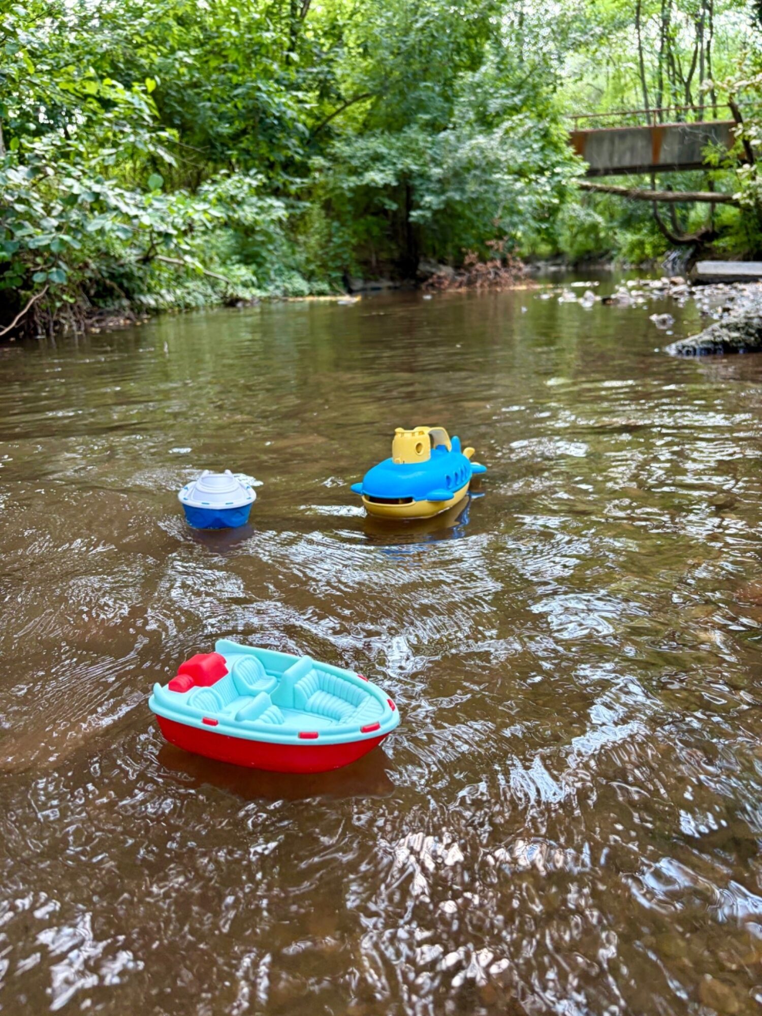 Three toy boats floating on a river with green trees and a wooden bridge in the background.