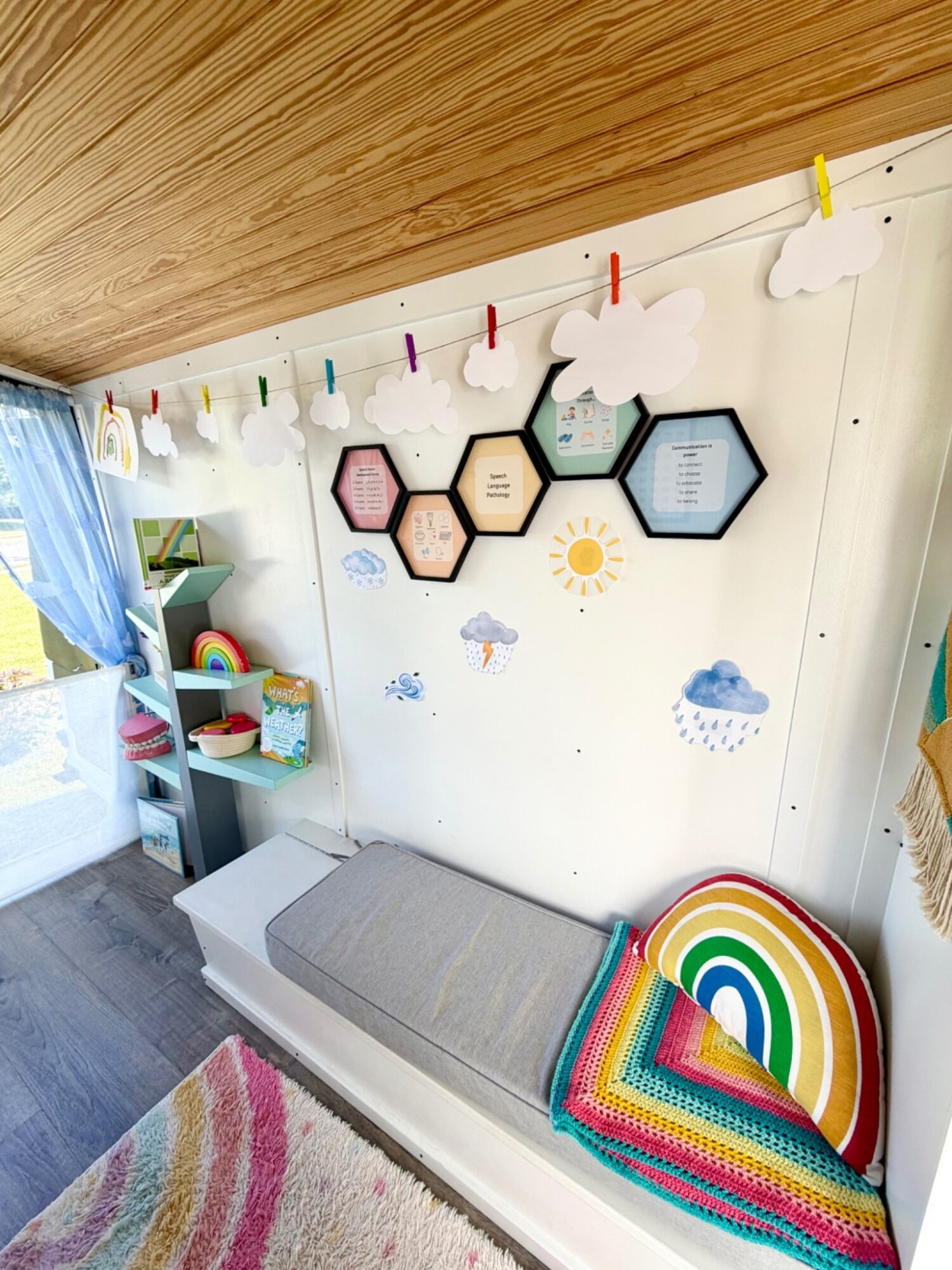 Child's room with a white wall decorated with cloud-shaped paper clips, hexagon-shaped wall art, and a rainbow pillow on a gray bench.