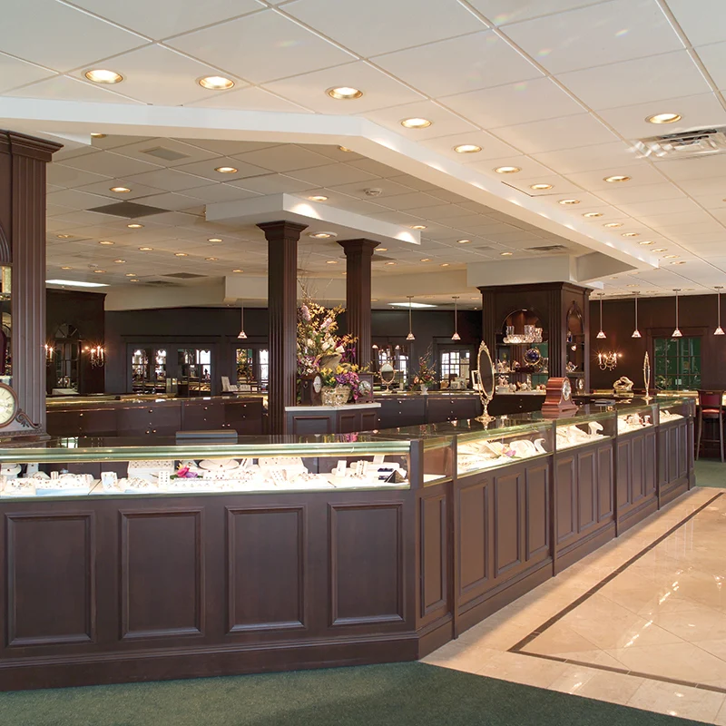 Jewelry display counter in a store with dark wood finish, glass cases, and ceiling lights, with a seating area in the background.