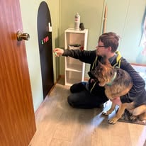 Person sitting on the floor with a dog, reaching towards a black door with a heart-shaped sticker, in a room with a small white shelf.