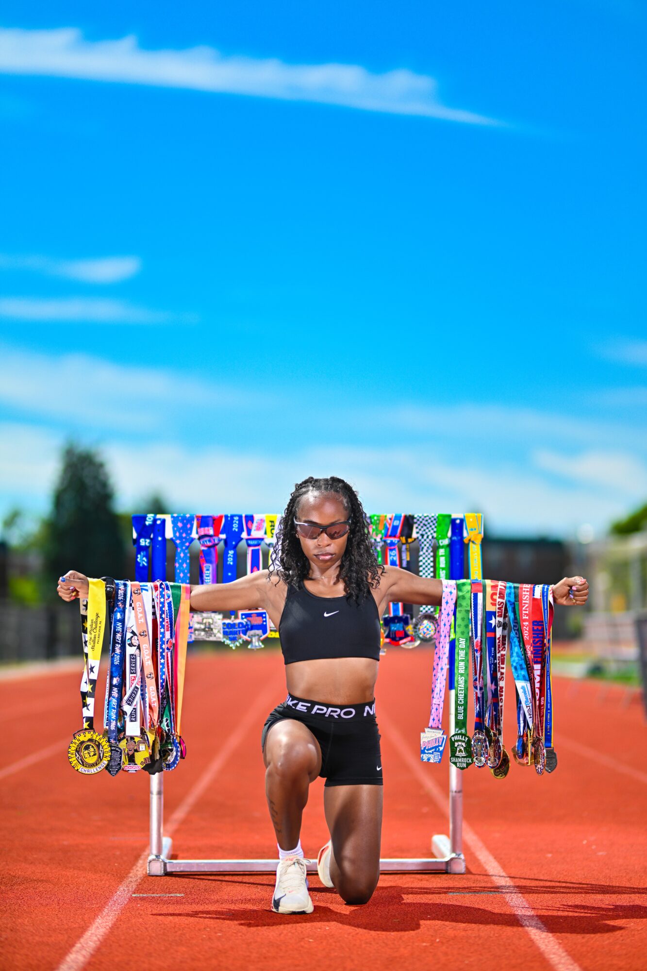 Young female athlete kneeling on a track with arms outstretched, holding numerous medals and ribbons, under a blue sky.