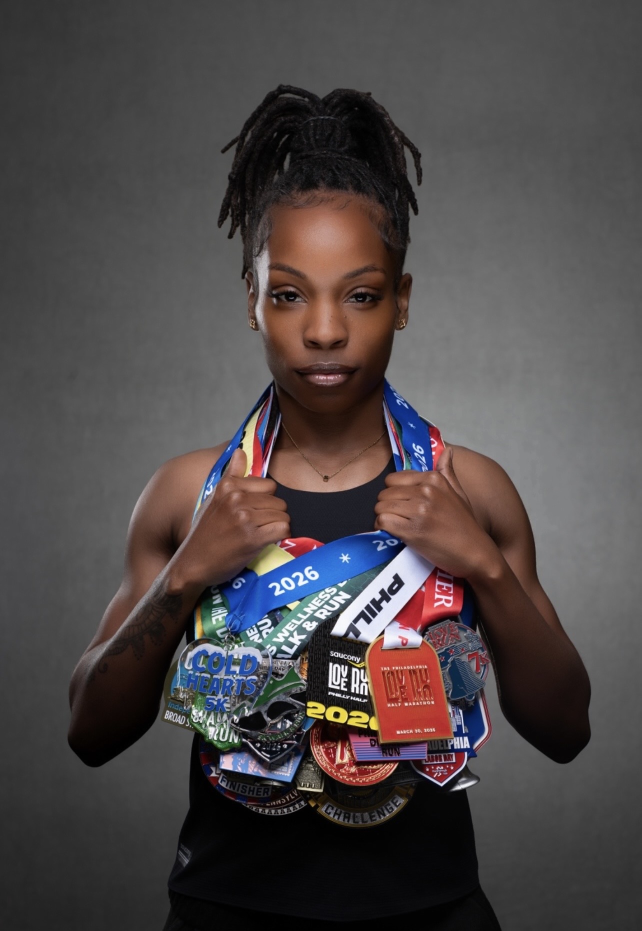 Young woman with braided hair holding multiple medals around her neck, looking confidently at the camera, gray background.