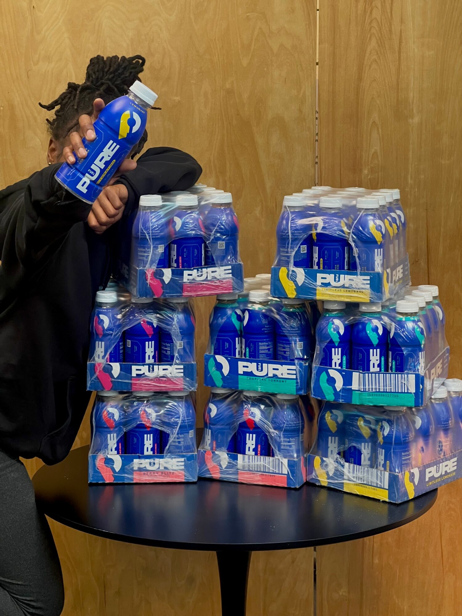 Child with dreadlocks holding a bottle of Pure bottled water, standing next to stacked packs of bottled water on a table.