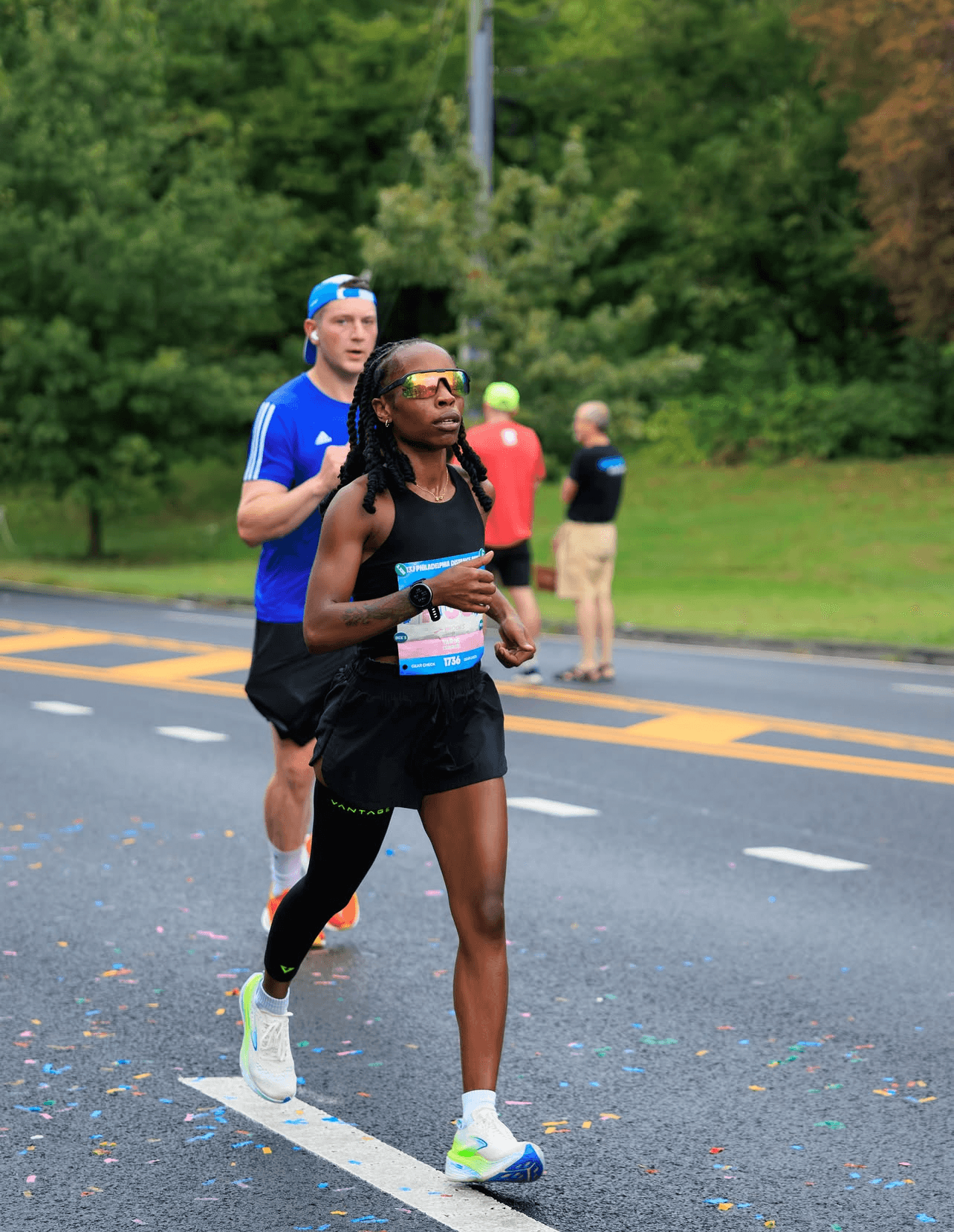 Two runners, a woman and a man, running on a road with trees in the background, woman in front wearing black shorts and tank top.