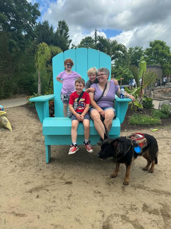 Three children and an adult sitting on a large turquoise outdoor chair, with a dog nearby, in a garden setting.