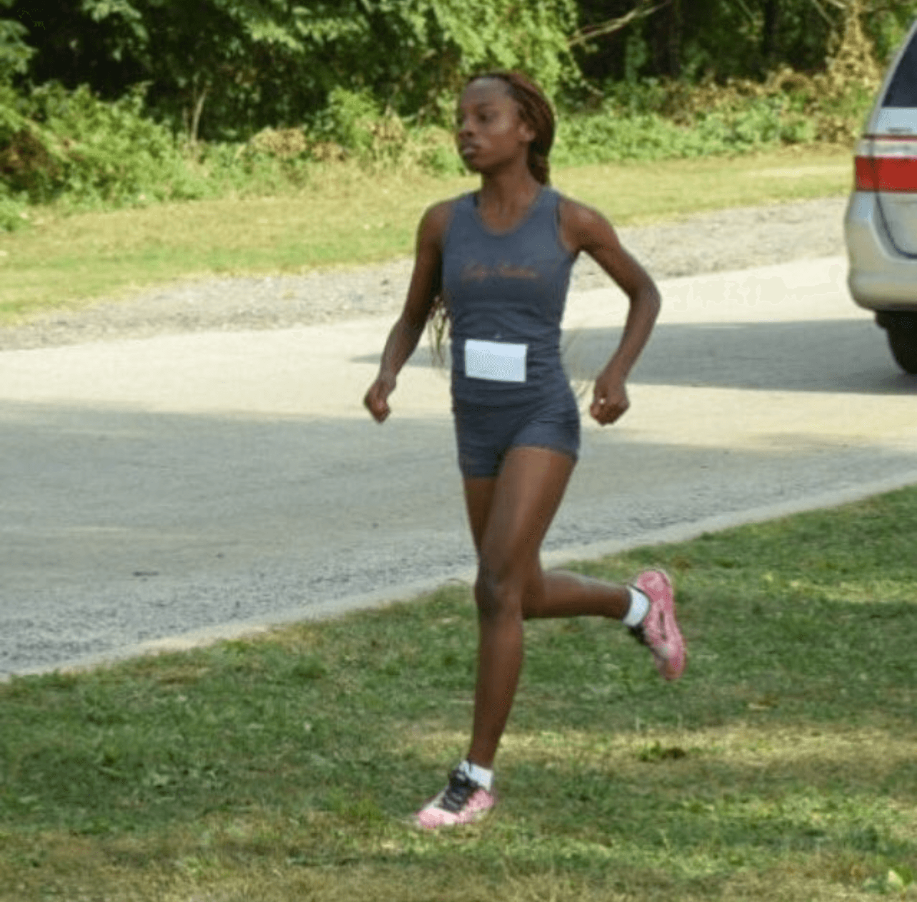 A woman running outdoors on grass and pavement, wearing a dark athletic outfit and pink running shoes.