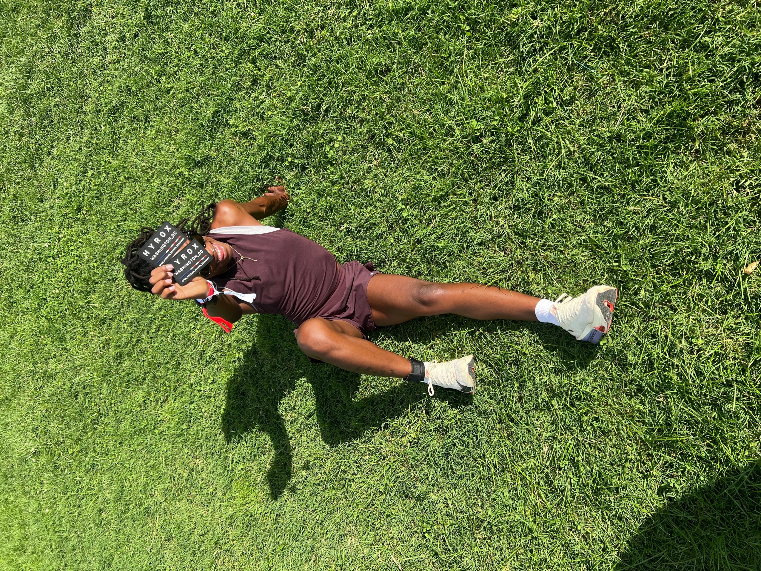 Person lying on grass holding a clapperboard, wearing a purple outfit and white shoes, shadow visible on grass.