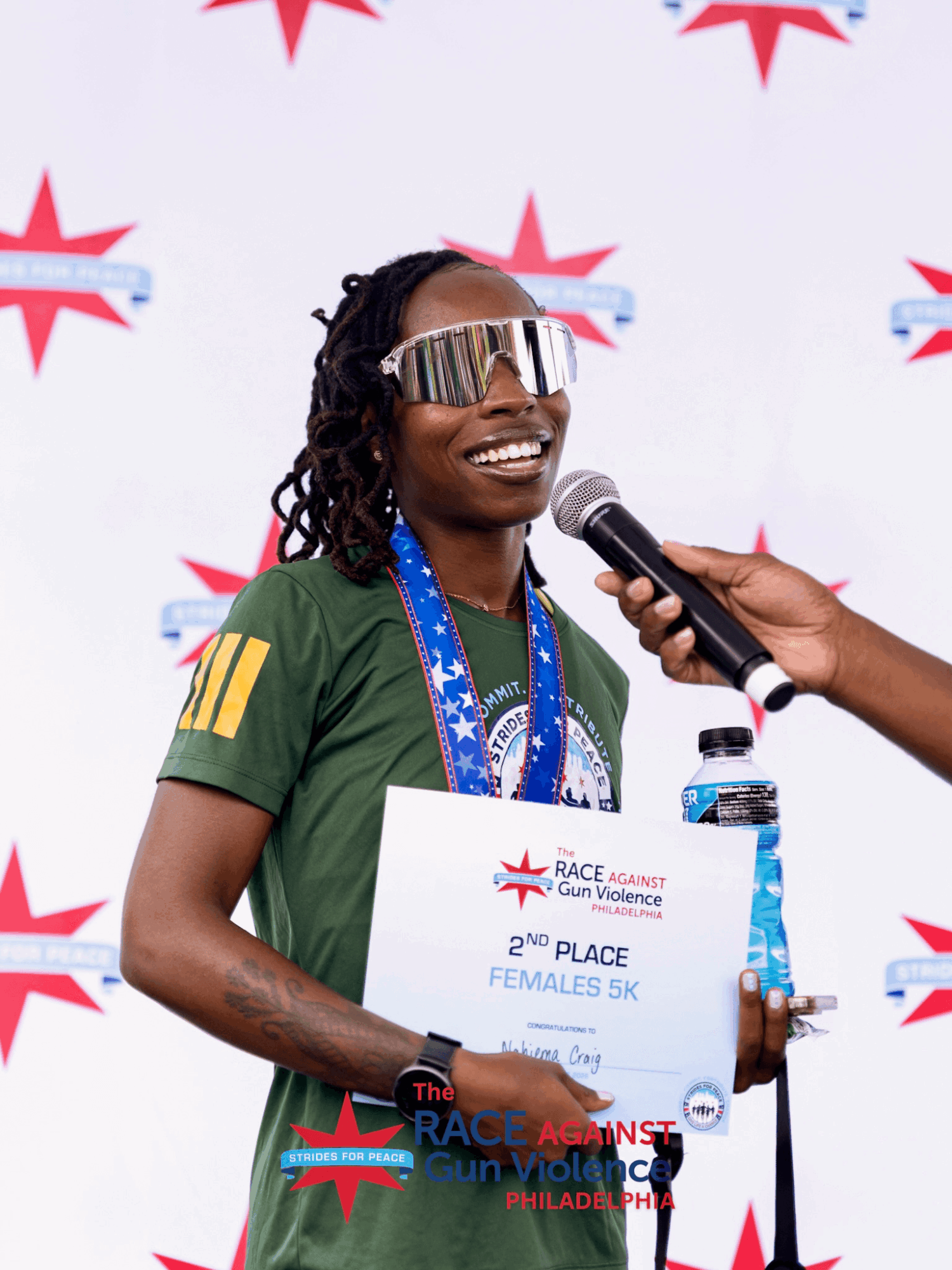 Smiling woman in green sportswear holding a certificate, wearing sunglasses, with a microphone held near her face, and a water bottle nearby.