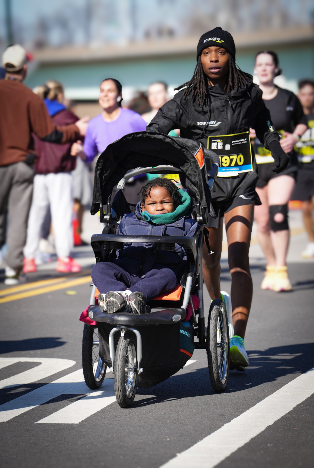 A woman pushing a stroller with a child during a race, with other runners and spectators in the background.