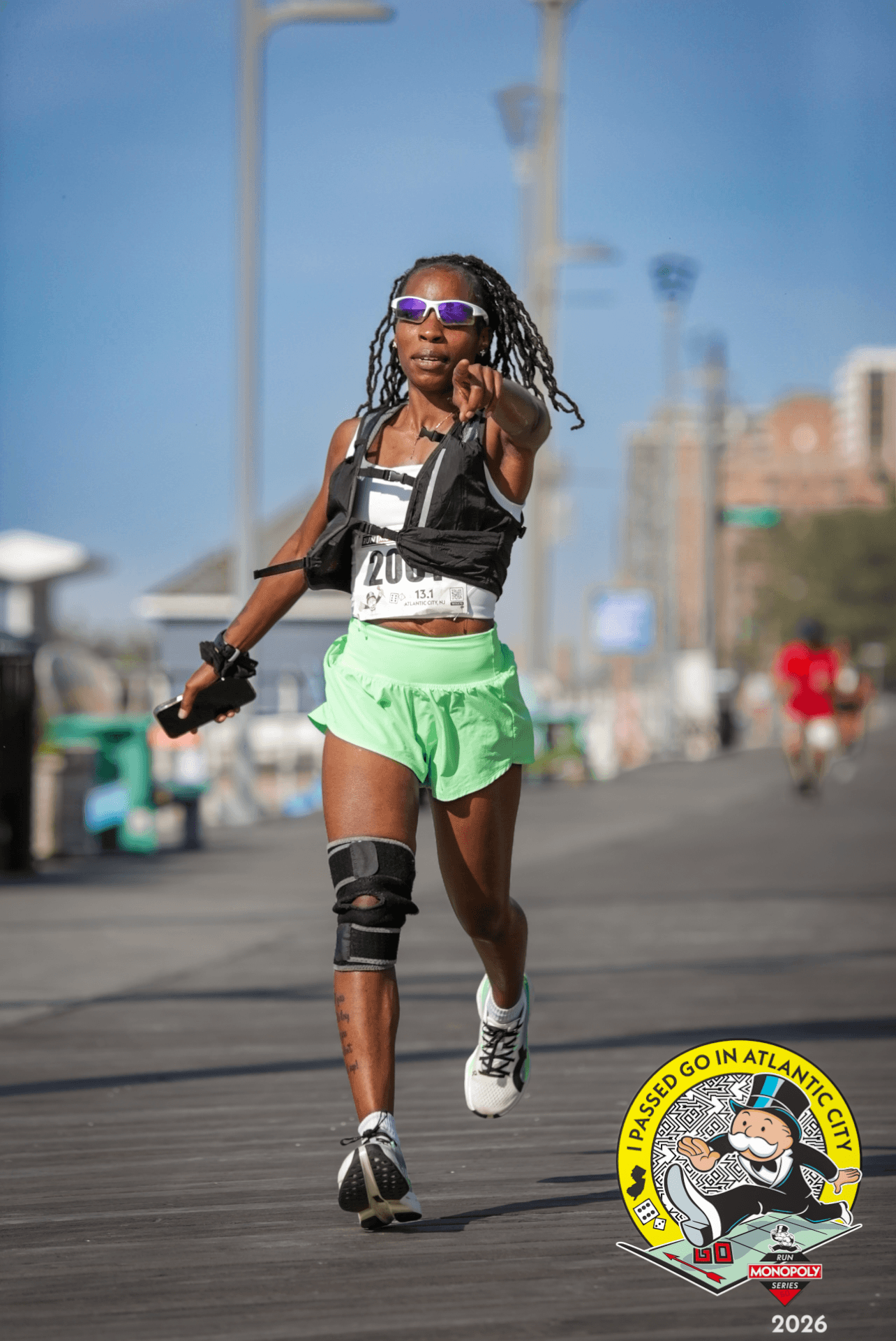 Female runner with sunglasses, wearing a black vest, green shorts, and a knee brace, running outdoors on a city street.