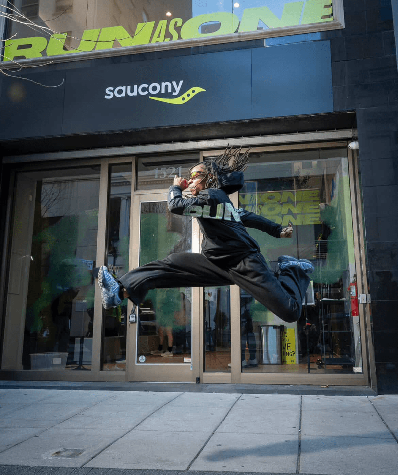 Person mid-air jumping in front of a store with Saucony and Run logo, hair flying, wearing athletic clothing and sneakers.