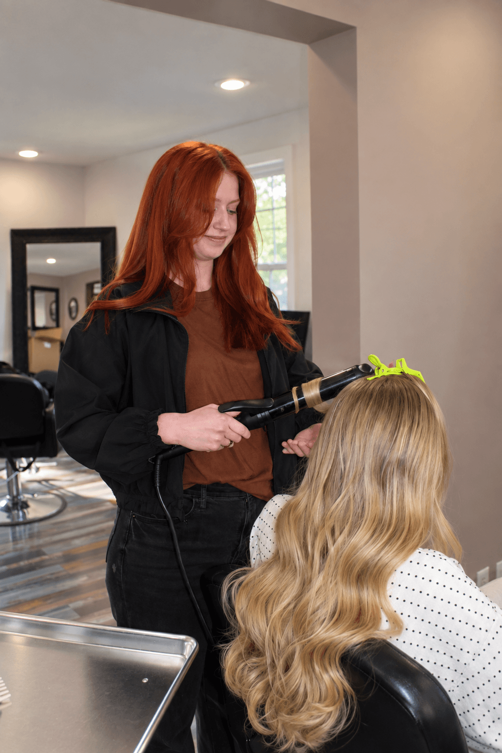 Hairdresser with long red hair styles a client's long, wavy blonde hair in a salon. Bright interior with mirror and window.