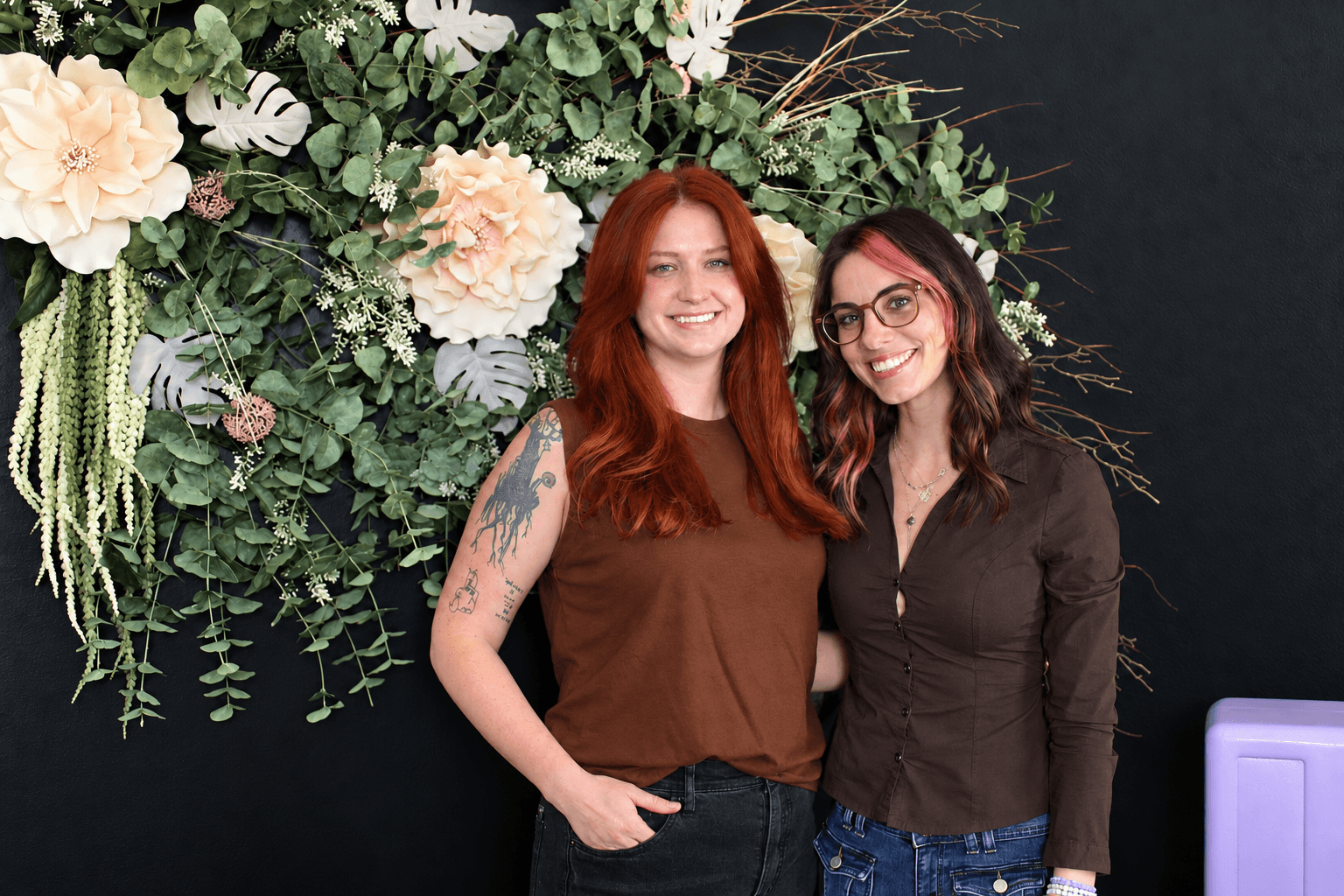 Two women smiling in front of a floral backdrop, one with red hair and the other with dark hair and glasses.