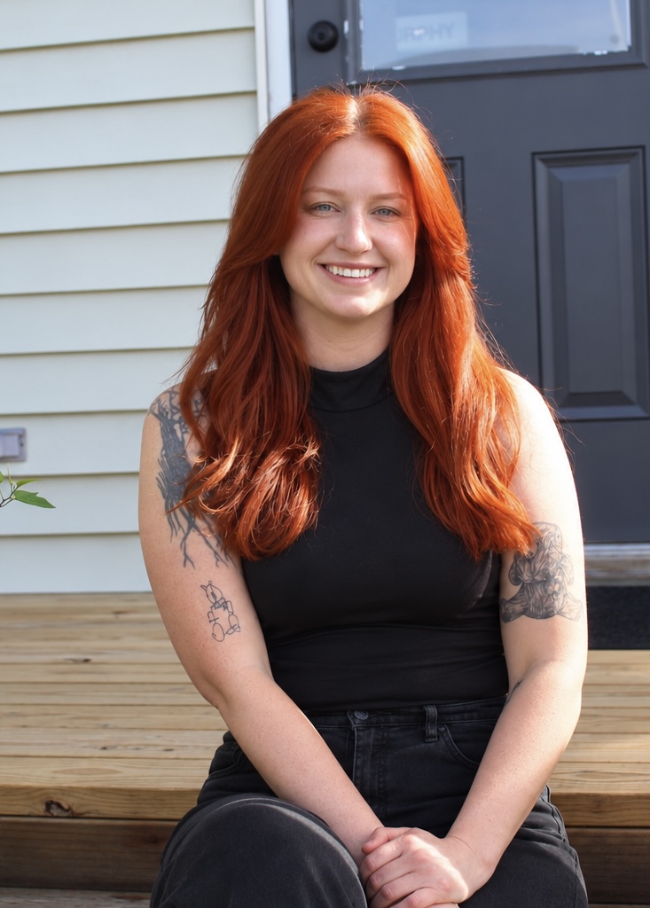 Young woman with long red hair smiling, sitting on wooden steps outside a house, wearing a black sleeveless top.