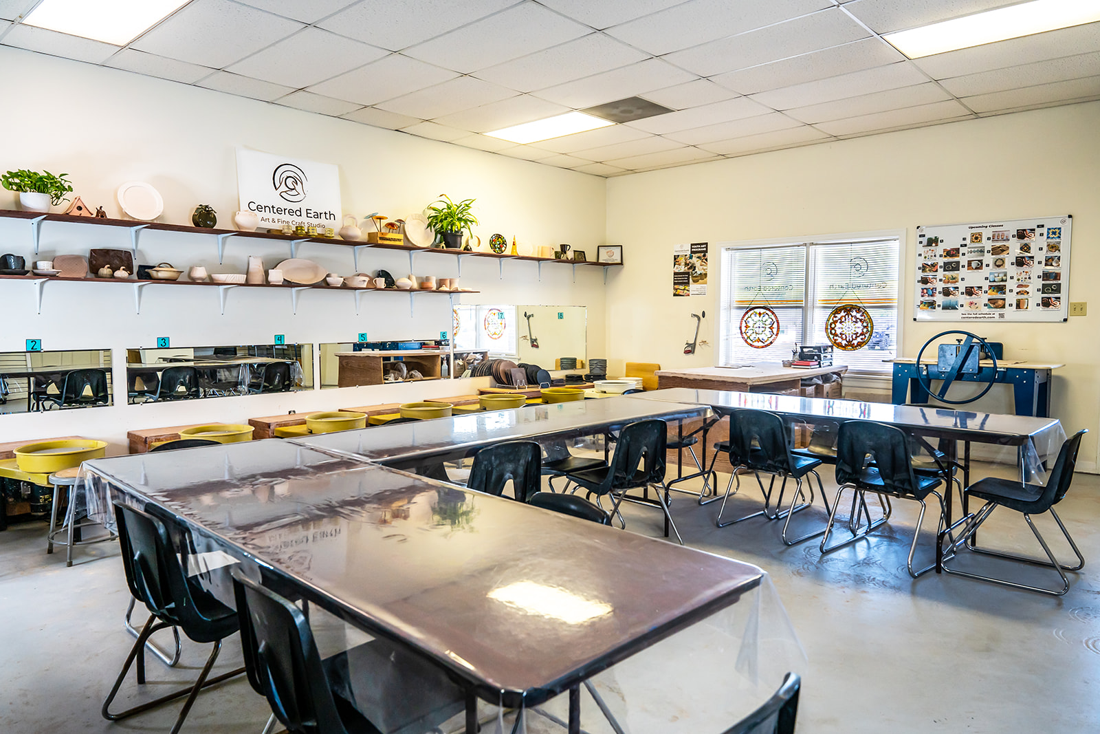 Classroom with tables, chairs, shelves, and windows, decorated with plants and educational materials.