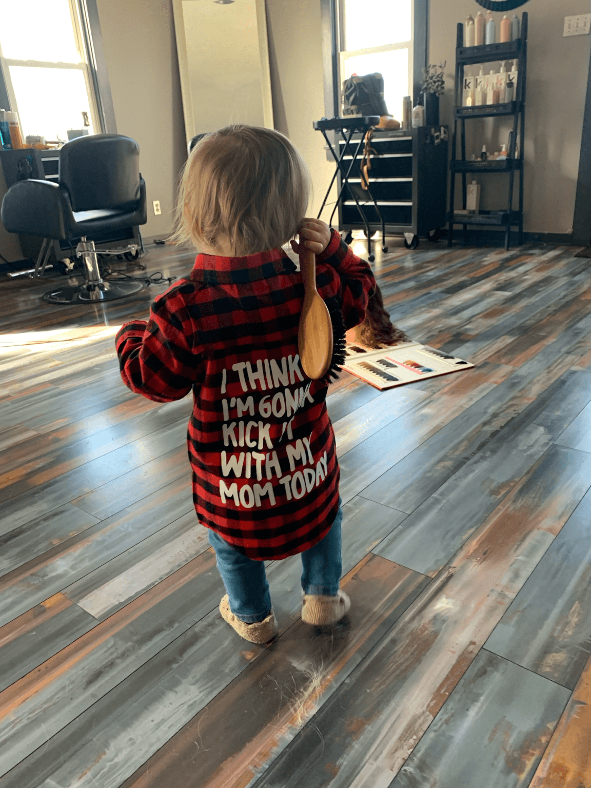 Child with blonde hair wearing a red and black striped shirt, holding a yellow object, standing in a room with wooden floor.