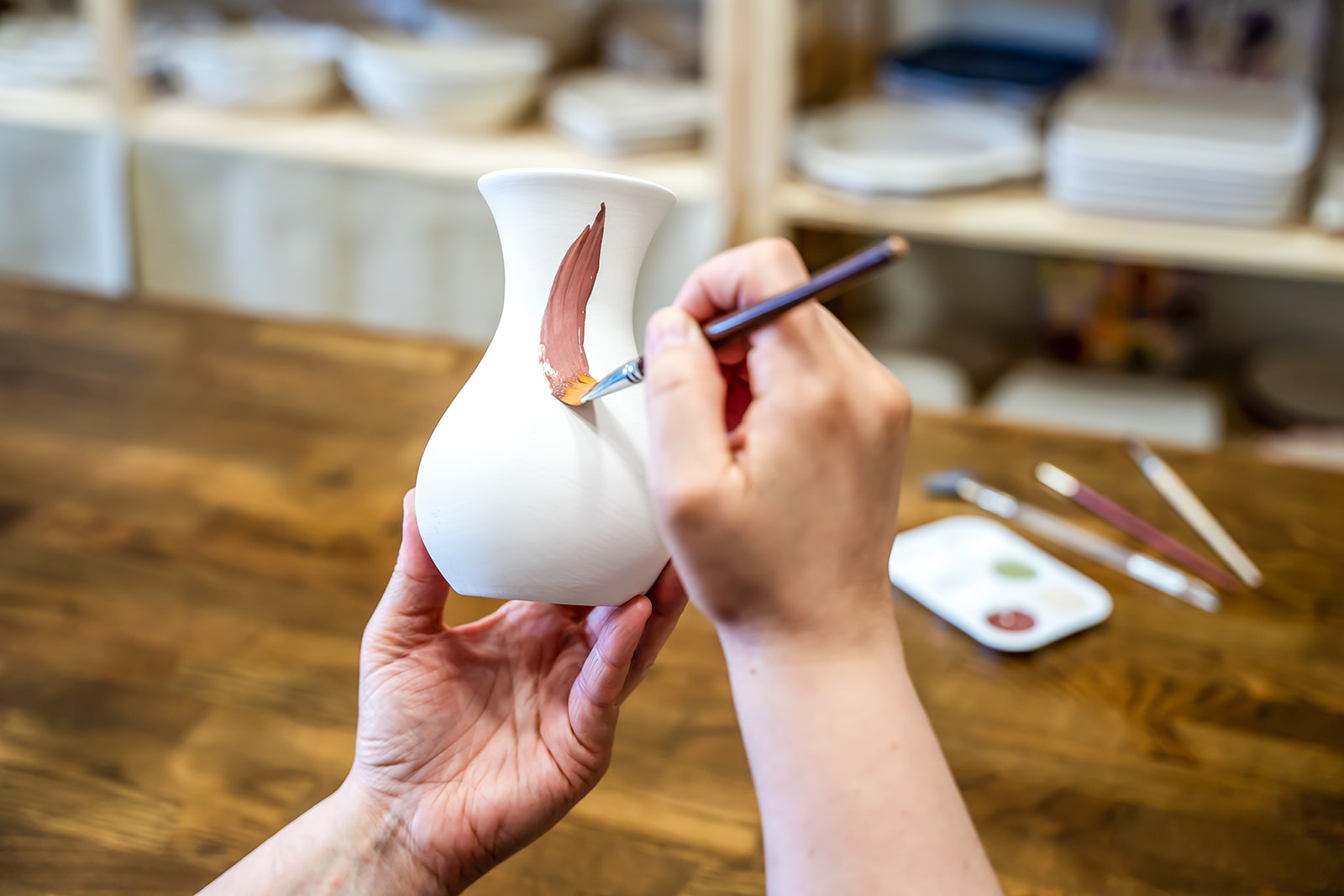 Person painting a white ceramic vase with a small brush, on a wooden table, with art supplies in the background.