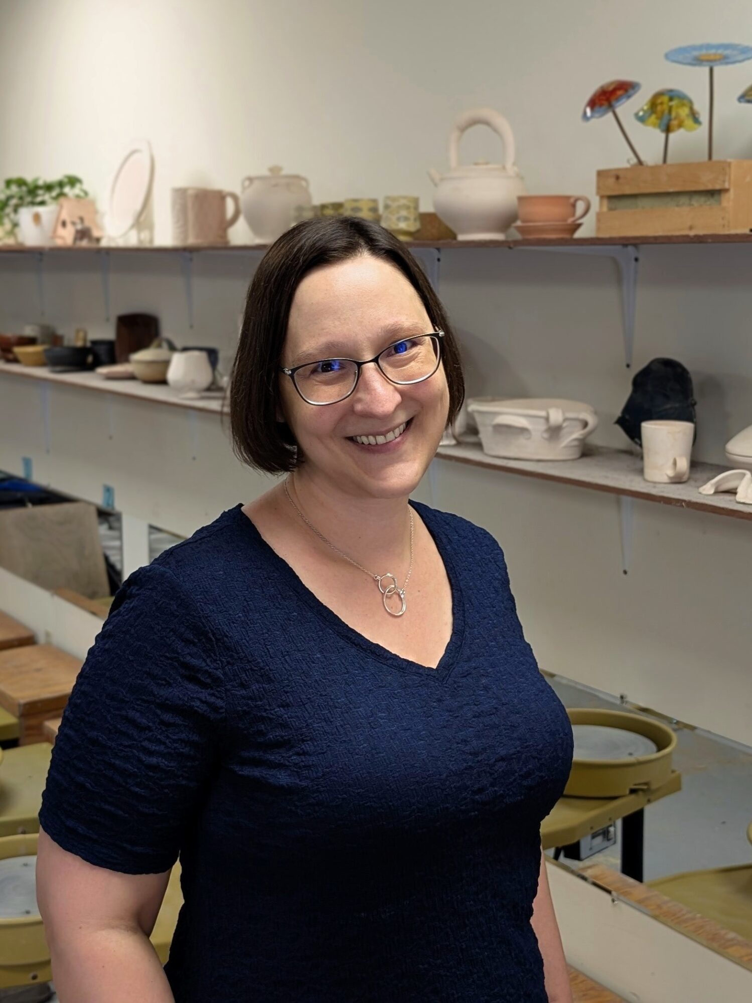 Smiling woman with glasses and short dark hair in a store with shelves of pottery and ceramics behind her.