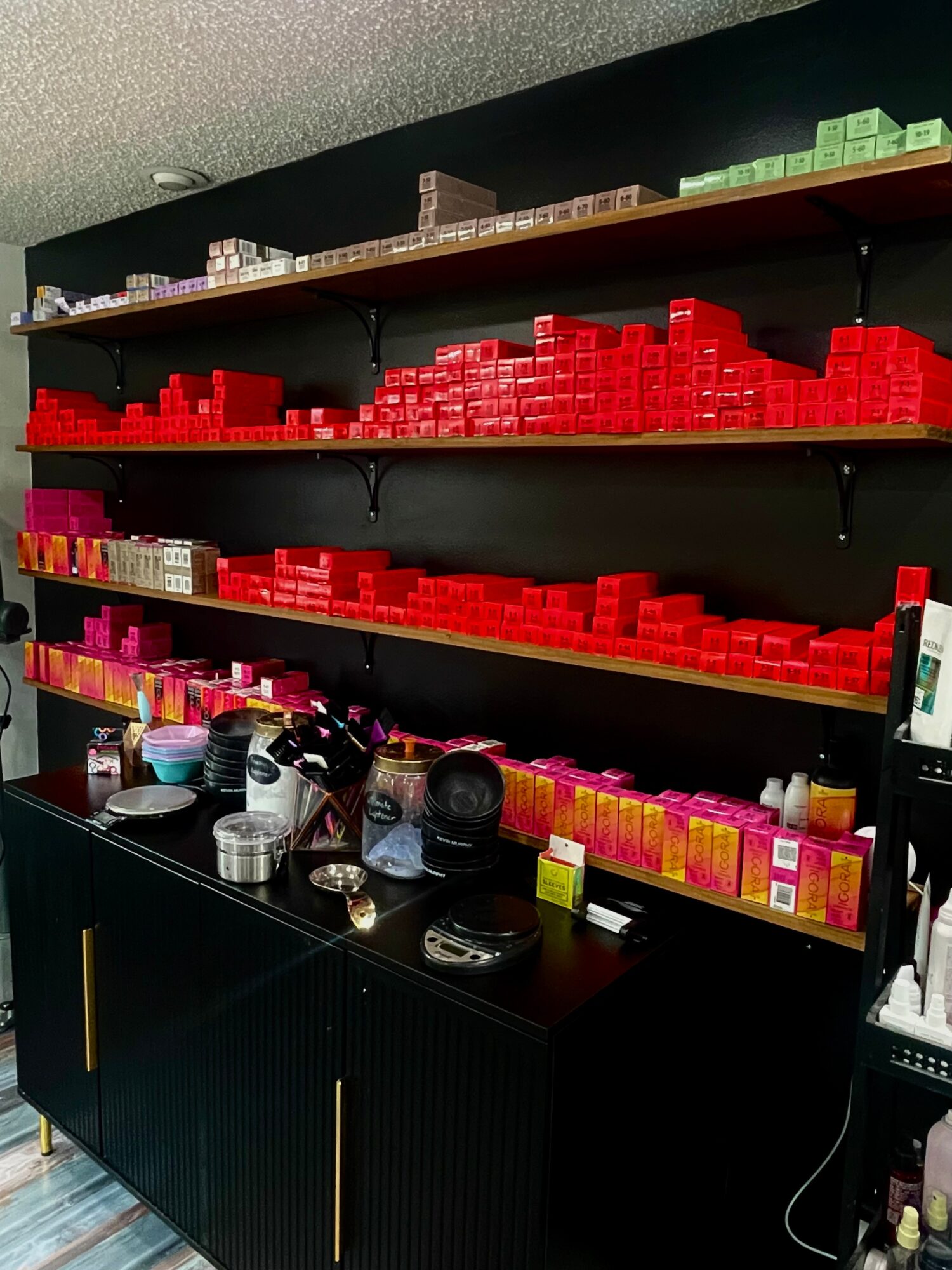 Shelves filled with red, pink, and brown boxes, with various items on a black cabinet below, against a black wall.