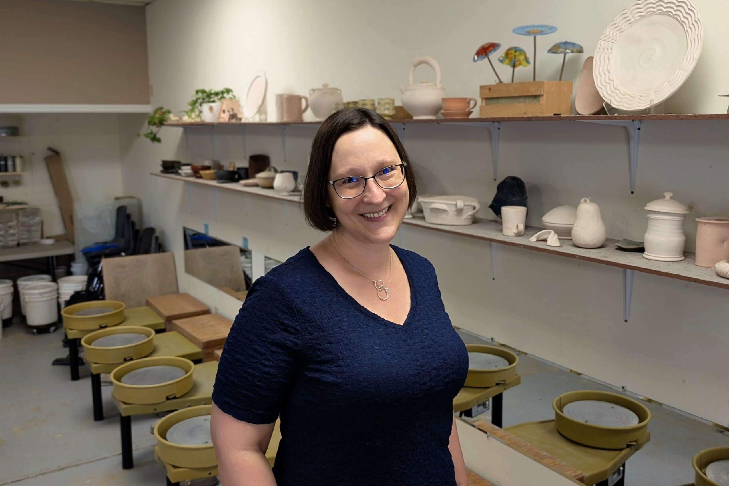 Woman with glasses and short dark hair smiling in a pottery studio with shelves of ceramics behind her.