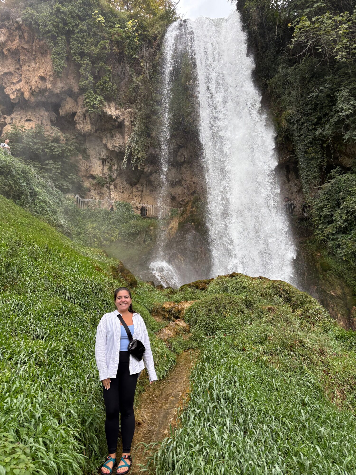 Person standing on grass near waterfall with trees in background, wearing white shirt and black pants.