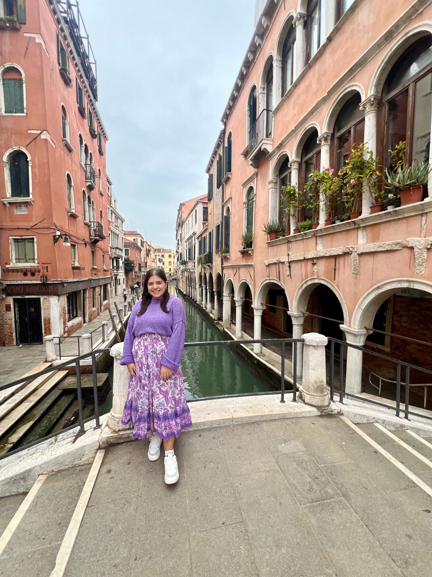 Young woman in purple dress and white shoes standing on a bridge over a canal with colorful buildings on both sides.