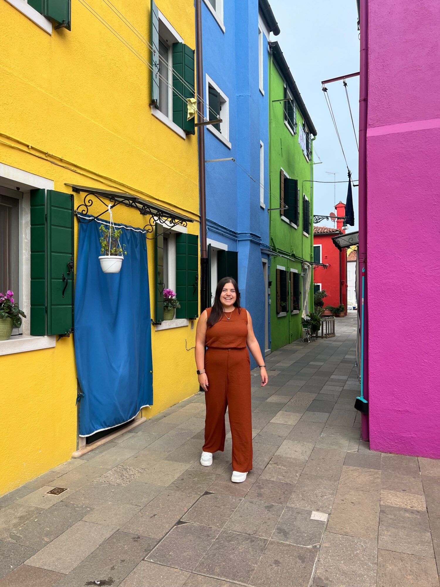 A woman stands on a sidewalk with colorful buildings in the background, wearing a brown outfit and smiling.