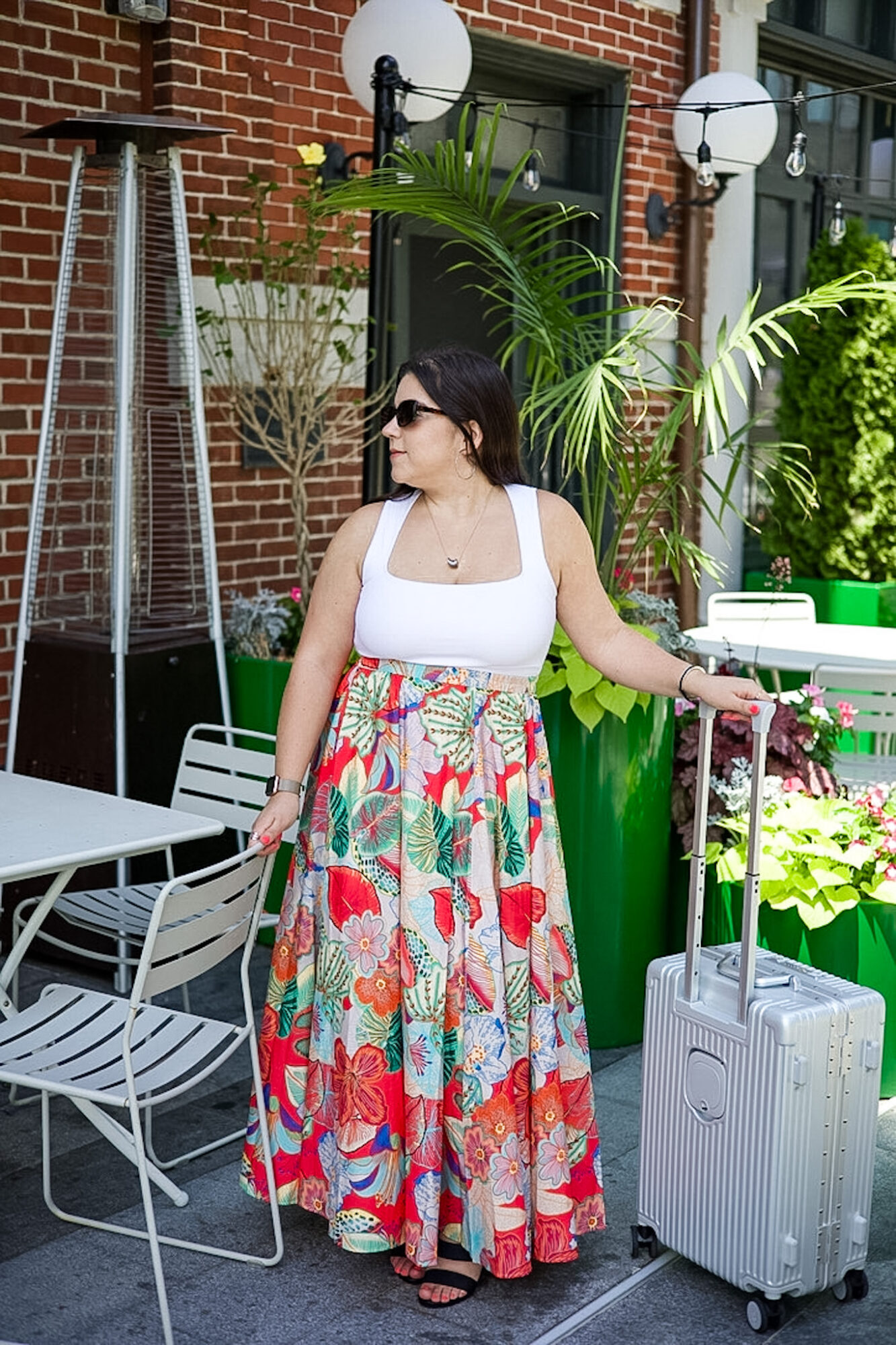 Woman in sunglasses wearing a white top and colorful floral skirt with a suitcase outdoors near plants and brick building.