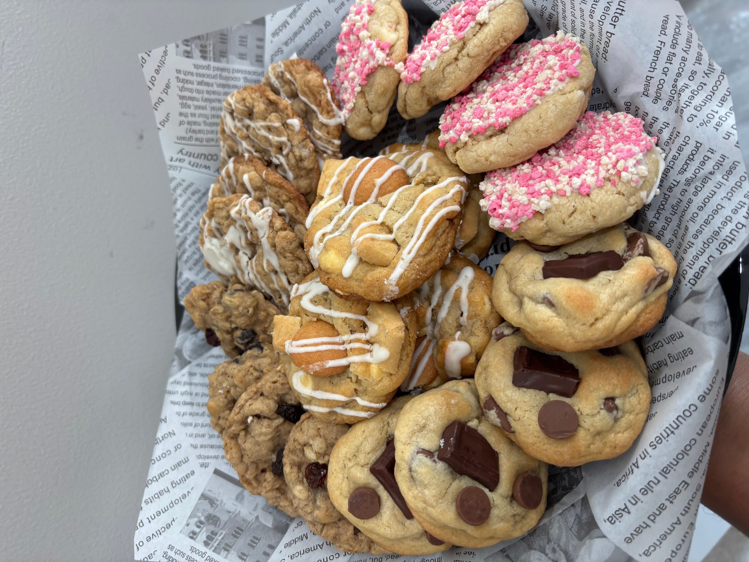 Assorted cookies and pastries on newspaper-lined tray, including pink-topped, drizzled, and chocolate chip cookies.