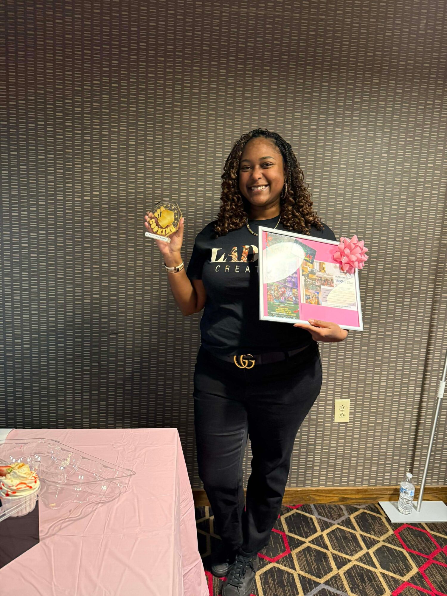 Woman smiling holding a colorful card and a small gift, standing near a pink table with a cake, against a textured wall.