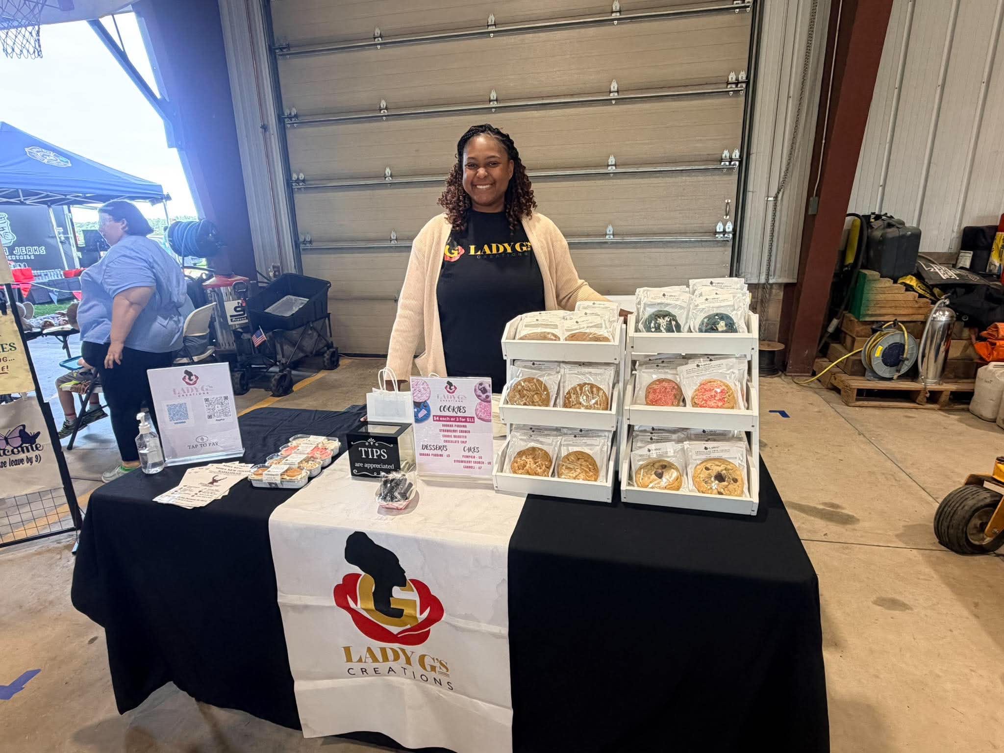 Woman standing behind a table with baked goods in a garage, smiling, with a Lady Lady logo on the tablecloth.