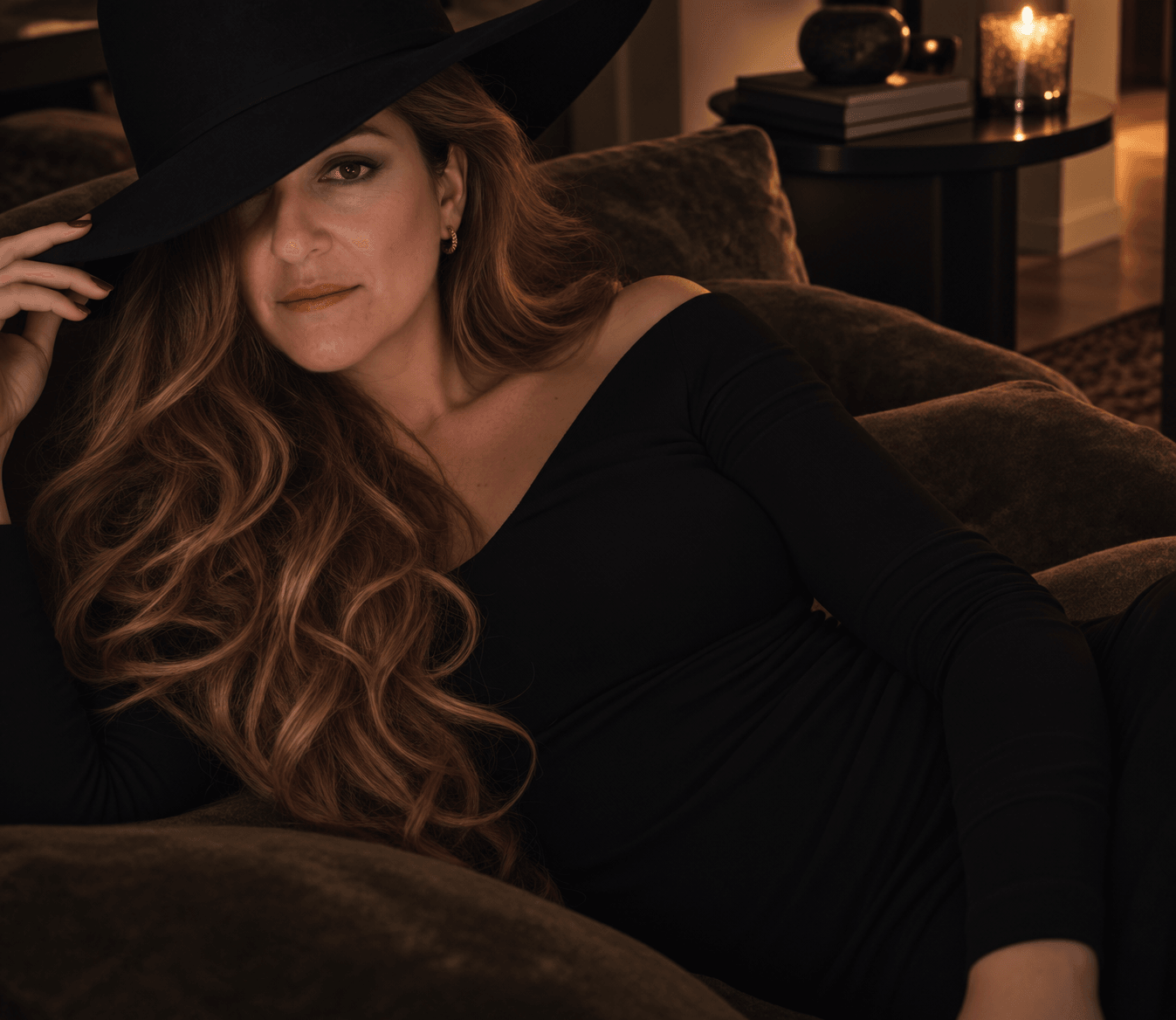 Woman with long wavy hair wearing a black dress and wide-brimmed hat, sitting on a sofa in a dimly lit room.