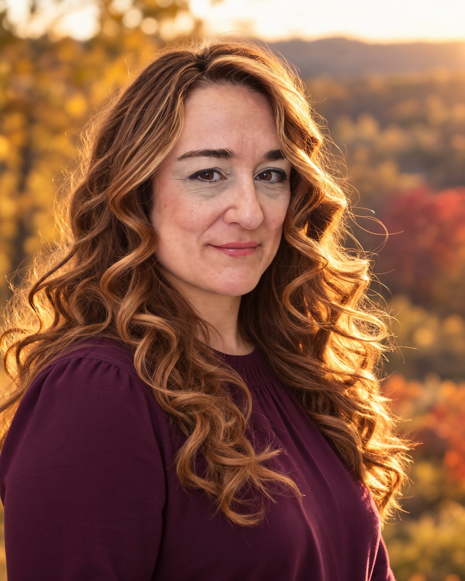 Woman with long, curly hair outdoors during sunset, wearing a purple top, with a blurred autumn landscape background.