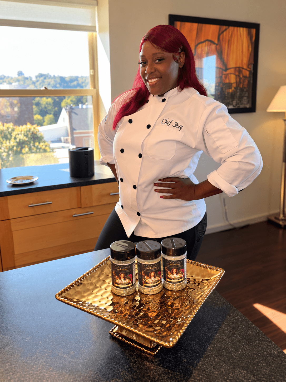 Woman in chef's coat standing behind a table with three jars on a gold tray, smiling, in a bright room with window and artwork.