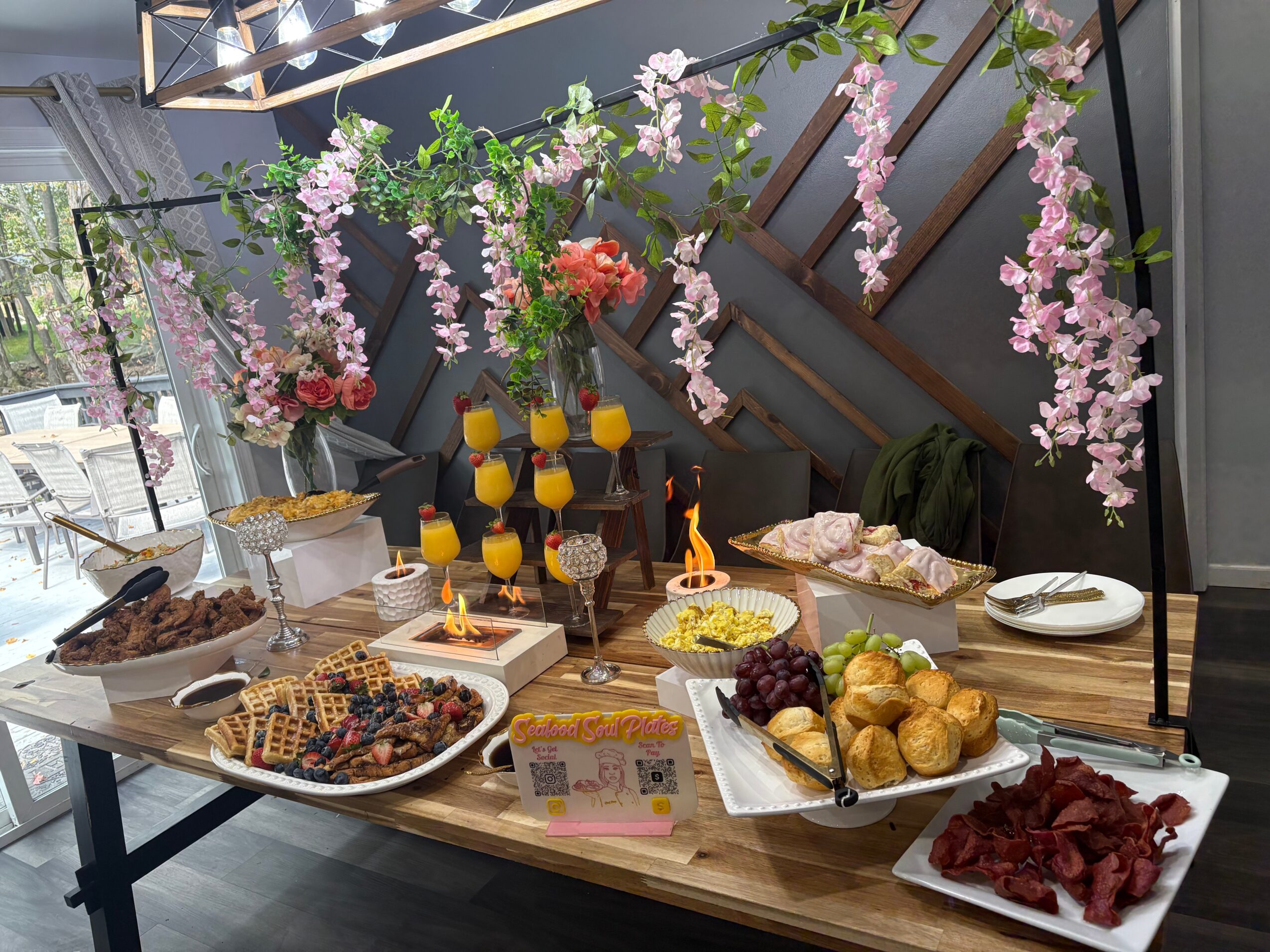 Table with desserts, fruits, and flowers, decorated with pink hanging blossoms and yellow candles, in a modern interior.