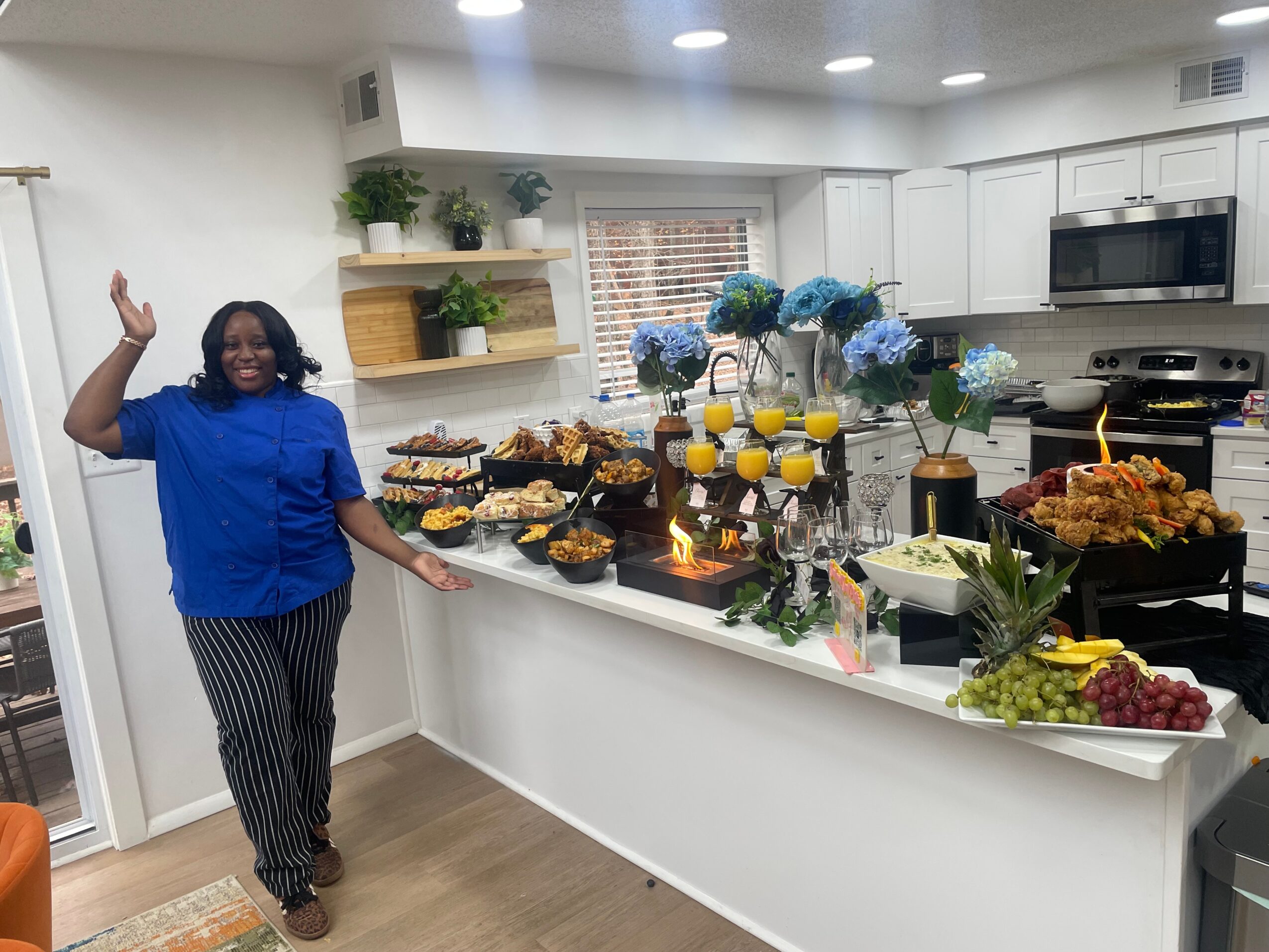 Woman in blue top and striped pants standing in a kitchen with a table of food and drinks, smiling and waving.