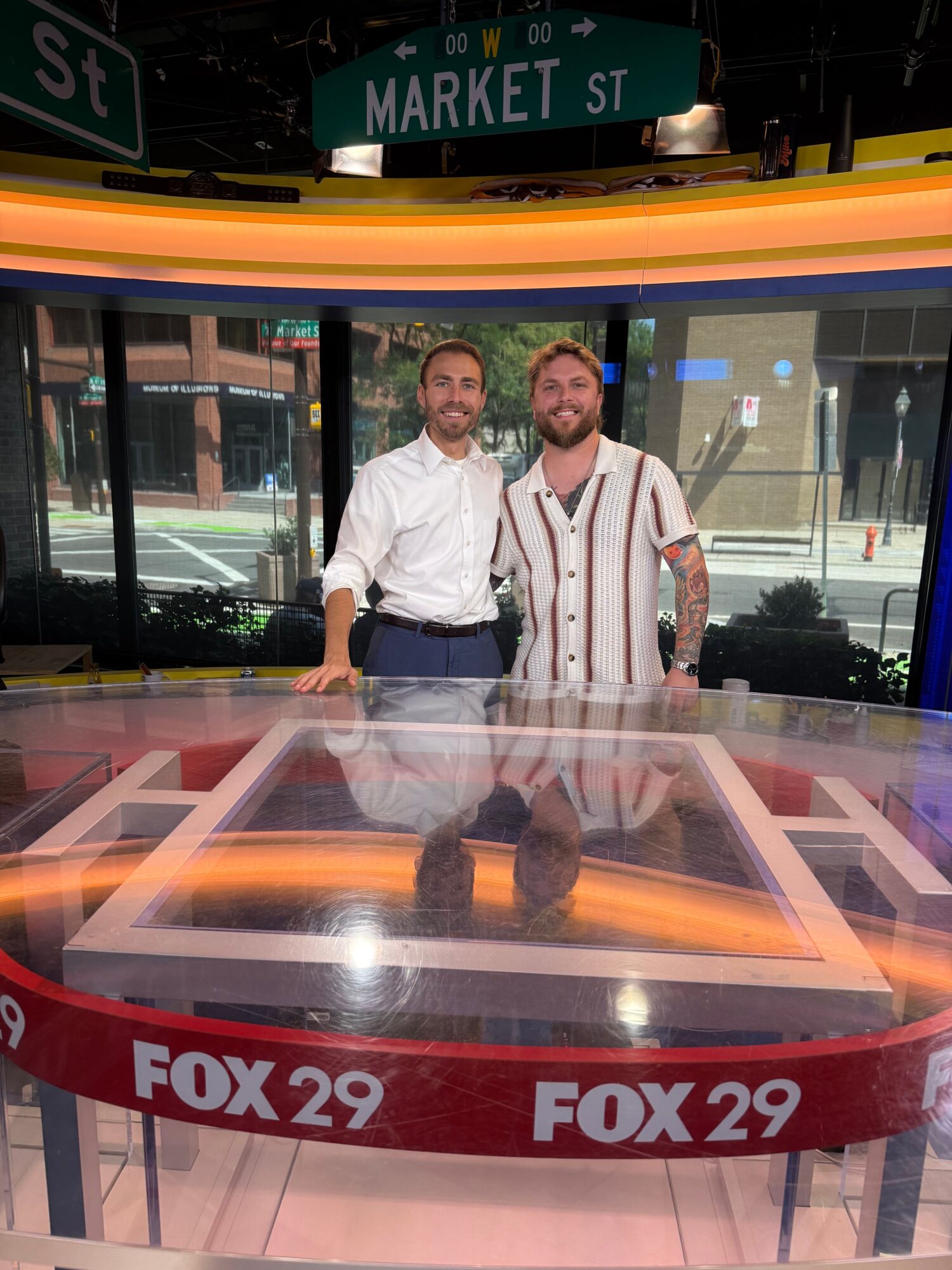 Two news anchors sit behind a glass desk in a television studio with FOX 29 branding, large windows, and cityscape background.
