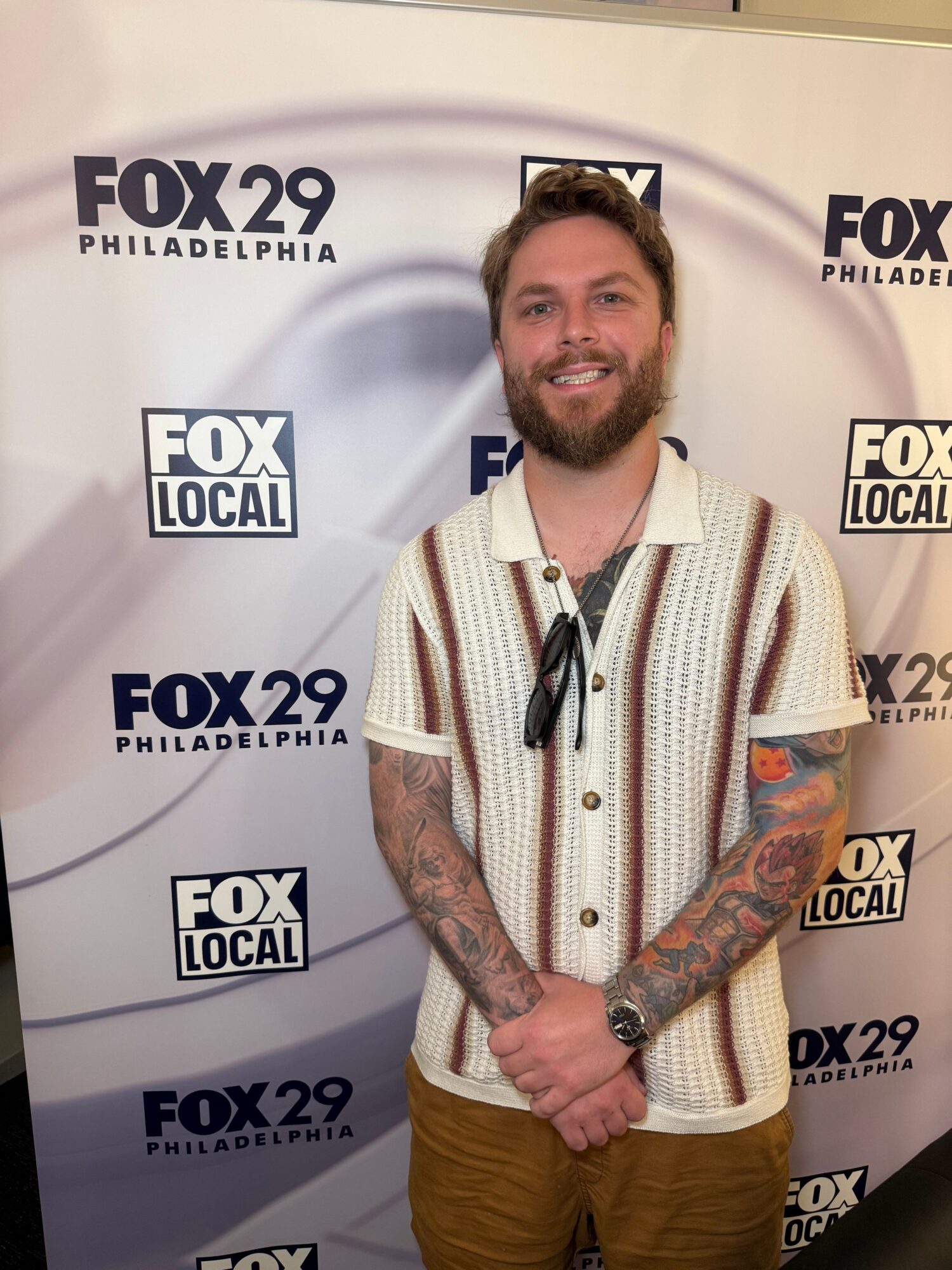 Man with tattoos and beard smiling, standing in front of FOX 29 Philadelphia backdrop, wearing a white striped shirt and brown pants.
