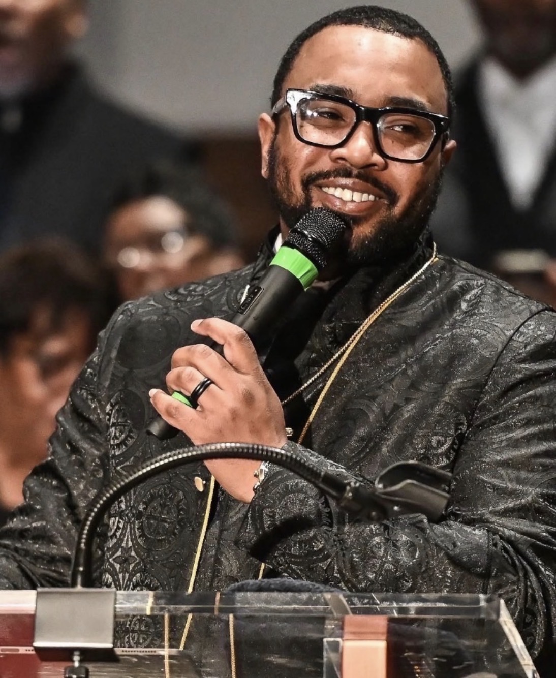 Man with glasses and a beard speaking into a microphone at a podium, smiling, wearing a dark patterned jacket, in a crowd.