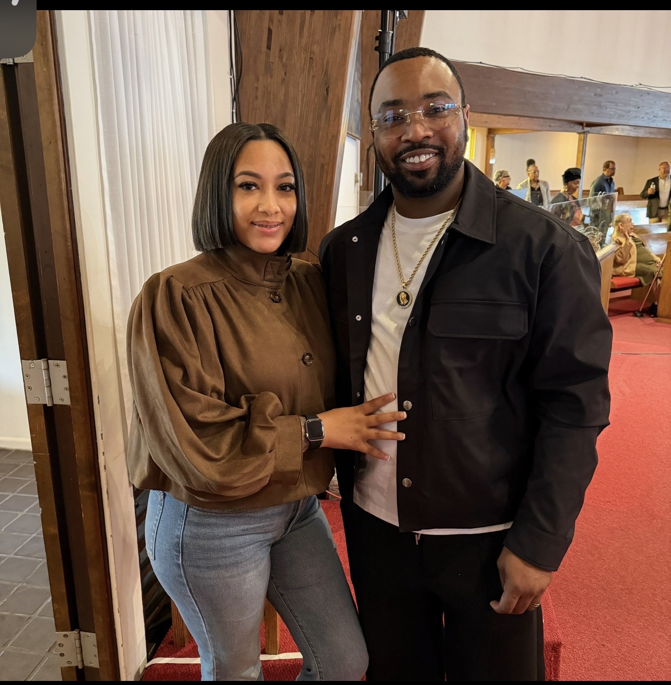 A woman and a man stand together indoors, smiling, with people in the background. The woman wears a brown top and jeans.