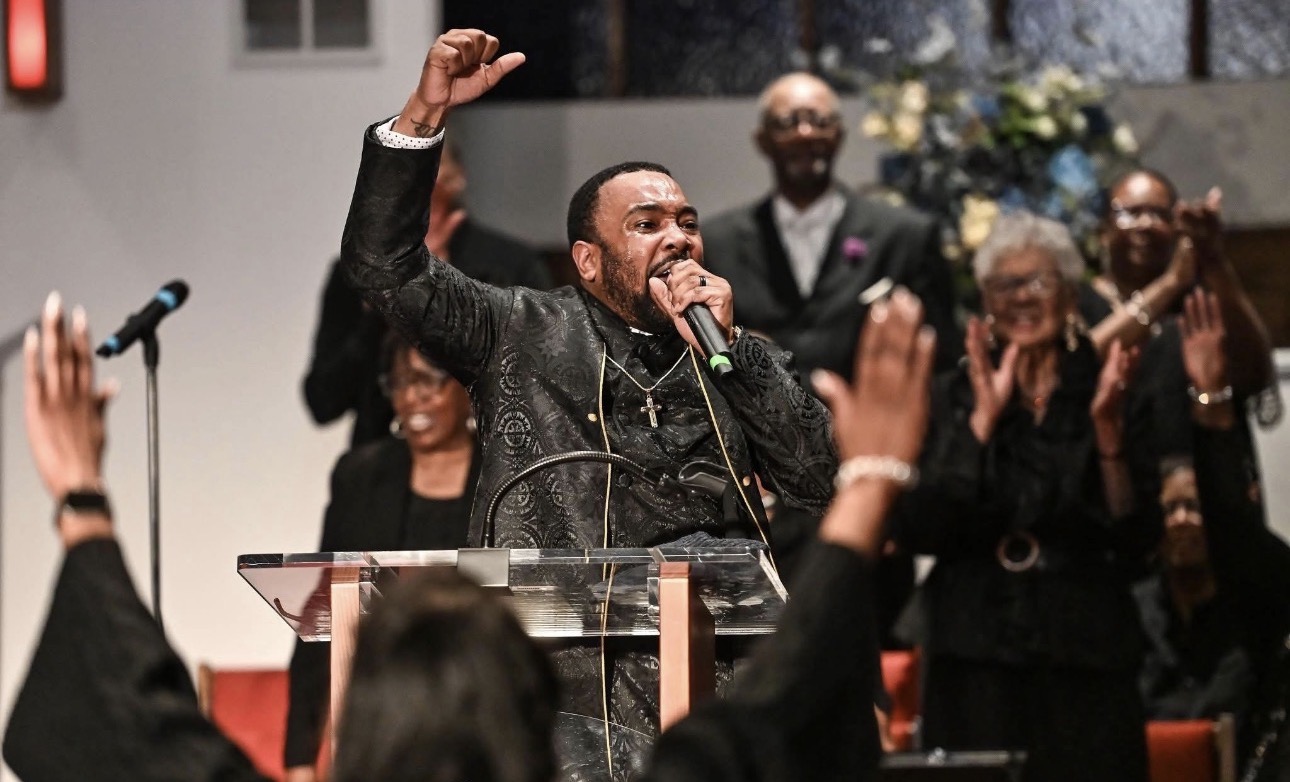 Man speaking passionately at a podium with raised hand, surrounded by smiling people clapping and celebrating.
