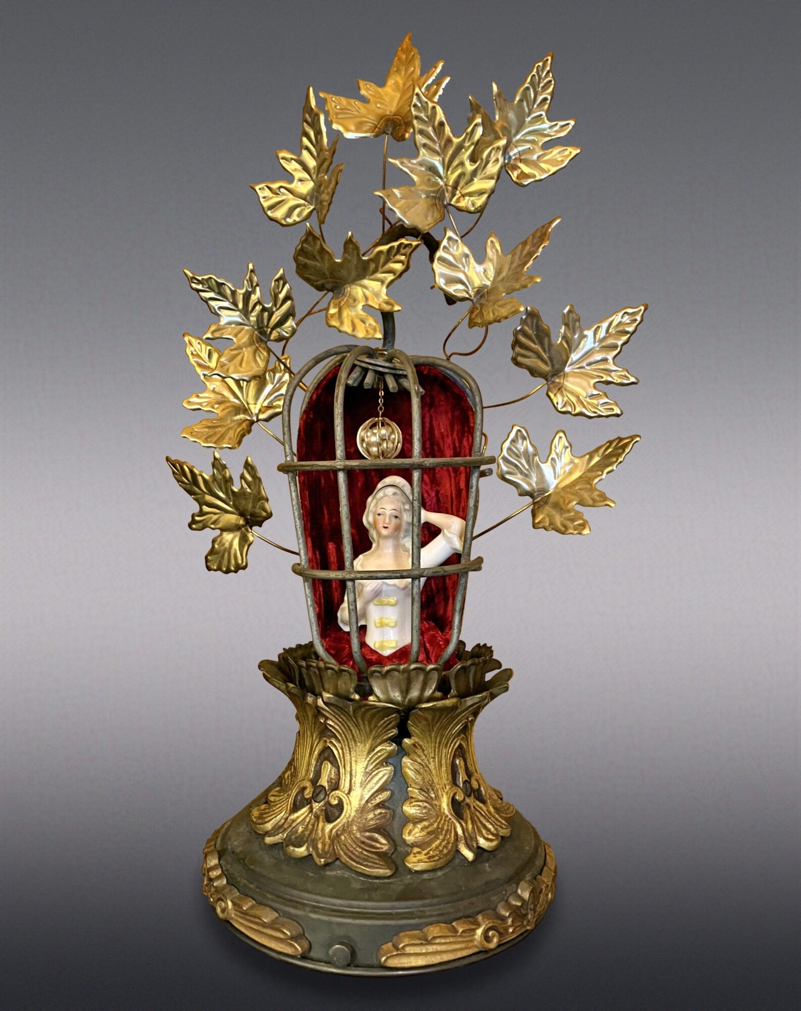 Decorative cage with a doll inside, surrounded by gold-colored leaves and butterflies, on a detailed base.