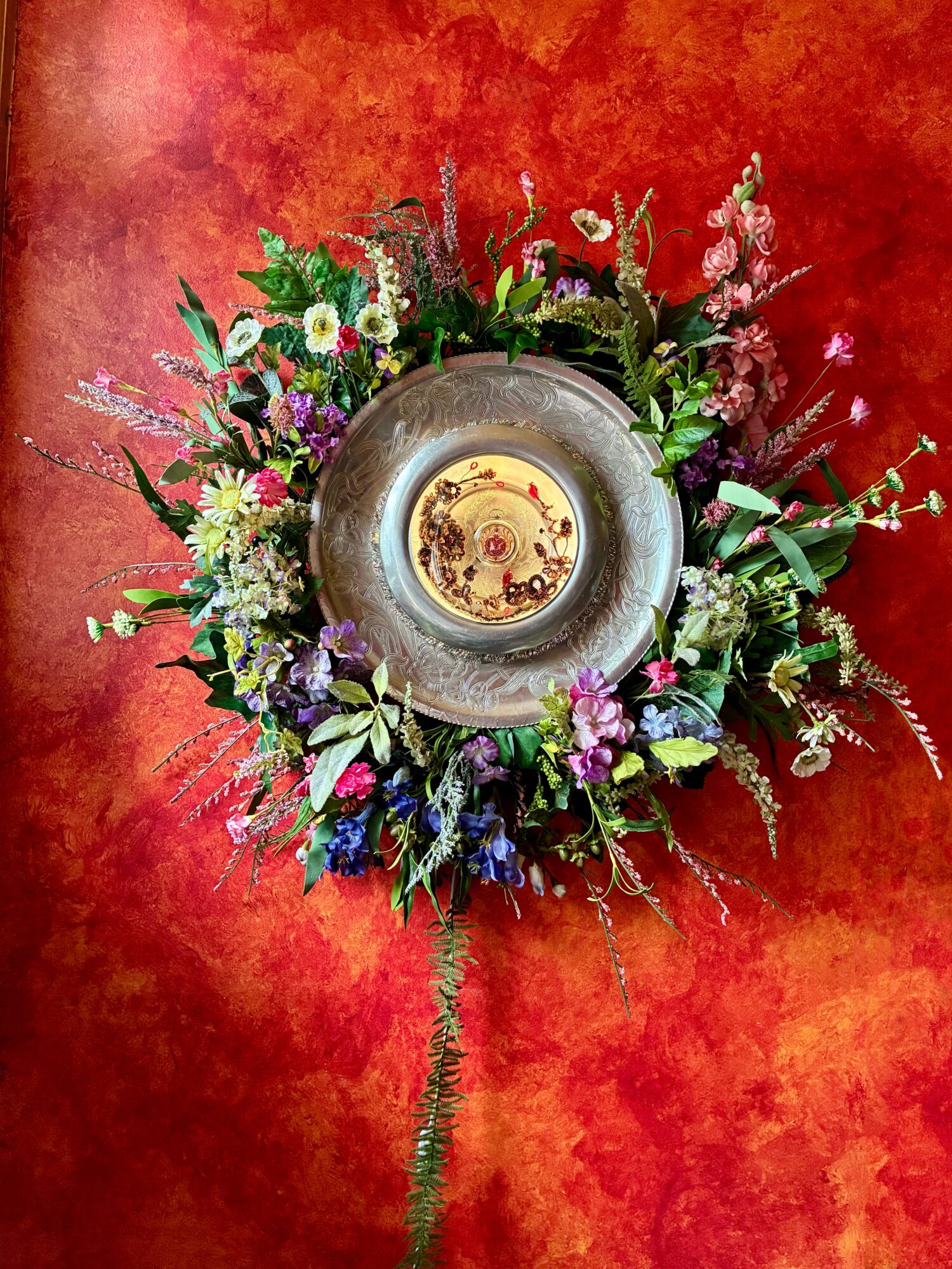 Top-down view of a circular arrangement of colorful flowers around a candle in a glass holder on a red surface.