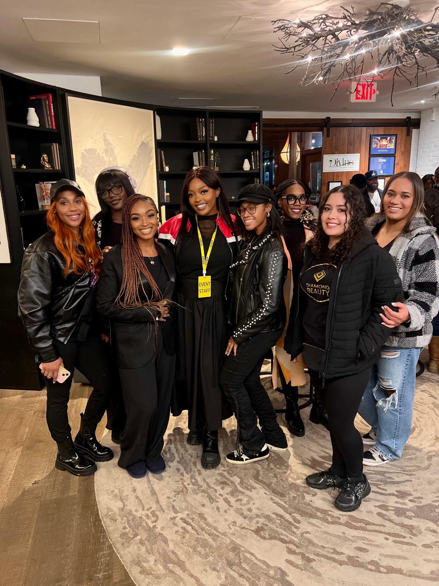Group of nine women standing together indoors, smiling, with bookshelves and a door in the background.