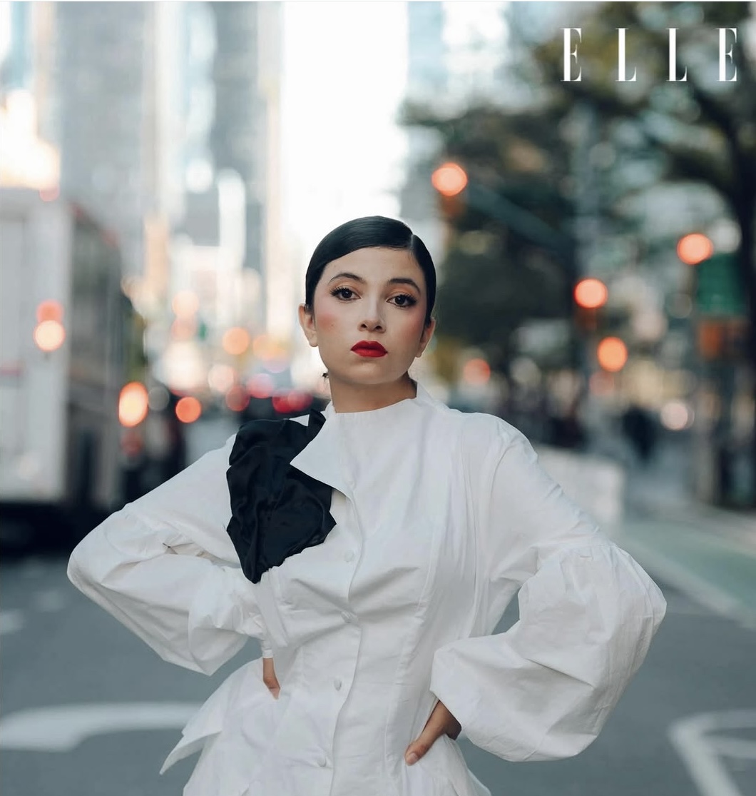 Woman with dark hair and red lipstick in a white blouse with puffed sleeves, standing outdoors with city background